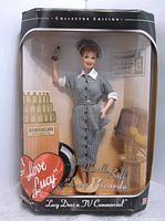 Front view of the 'I Love Lucy' Vitameatavegamin doll in its original packaging, showing doll in grey checkered dress, with display props and collector edition label.