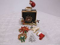 Eight Christmas themed brooches displayed with a round trinket box with Santa figure lid on top of a white surface.