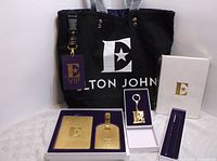 Full set displayed including black tote bag with white Elton John logo, VIP lanyard with badge, gold-tone passport holder and luggage tag, keychain, journal and pen, all new and in presentation box.