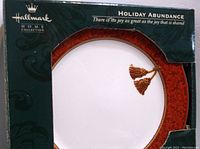 Close-up photo of the dinner plate in original packaging showing the Holiday Abundance design with red and gold border and gold tassels.
