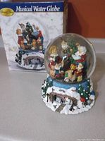 Overall view of the musical water globe with its original box in the background, showing the base decorated with snow and icicles and the figurines inside the globe.