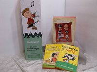 Full view of the lot showing the Musical Charlie Brown Christmas Tree box standing upright with 'The Original Musical Charlie Brown Christmas Tree' text, two vintage Peanuts books titled 'The Peanut' and 'Don't Bet on It Snoopy,' and the Hallmark Keepsake ornament box with Peanuts characters.
