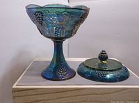 Blue carnival glass compote with grape vine pattern and detached lid shown on wood surface.