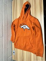 Orange youth hoodie with Denver Broncos logo on front, displayed hanging against wooden and brick background.