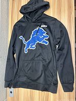 Black hoodie showing large blue lion logo on front with 'LIONS' near neckline, laying flat on a wood surface.