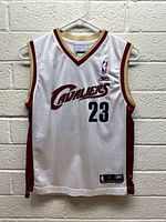 Front view of white Cleveland Cavaliers basketball jersey with number 23 and Cavaliers logo, hung against a brick wall.