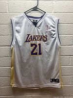 Full front view of white Los Angeles Lakers basketball jersey size Youth XL hanging on hanger against concrete background. Shows 'LAKERS' text and number '21', NBA and Reebok logos.