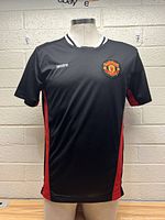 Front view of a black Manchester United soccer jersey on mannequin. Red side stripes and Manchester United crest on chest visible.