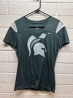 Front view of green women's T-shirt with silver Spartan helmet logo and light grey shoulder accents displayed on a hanger against a wall.