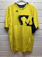 Front view of yellow Michigan Wolverines Adidas shirt hanging on a white hanger against white brick wall; shows large faded 'M' logo in navy and Adidas logo on chest.