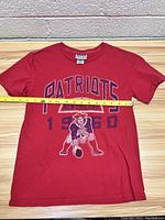 Full front view of red New England Patriots youth shirt with graphic of football player and text 'Patriots 1960 New England'.