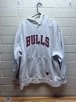 White Nike Chicago Bulls pullover hoodie displayed hanging against a wall, showing large 'BULLS' text in red with black outline on the chest and Nike swoosh above.