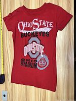 Red Ohio State Buckeyes women's T-shirt laid flat on wooden surface with shirt design visible.