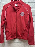 Front view of red fleece jacket with Ohio State Buckeyes embroidered logo on chest, hanging on white plastic hanger against a brick wall.