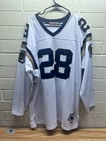 White Mitchell and Ness Marshall Faulk St Louis Rams jersey, number 28, hanging showing front and sleeves with team logos and numbers.