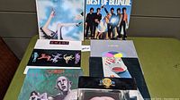 Vinyl records displayed on green surface including Queen, Heart, Yes, Kool & The Gang