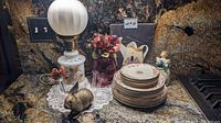 Photo shows vintage lamp with frosted glass globe on floral base, brass rabbit figurine, purple glass test tube rose vase with artificial flowers, Edwin Knowles china stacked plates with floral pattern, ceramic teapot light switch cover, all arranged on granite countertop.
