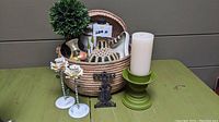 Large woven basket containing macrame wine bottle cover, ceramic pitcher vase, green candle holder with white candle, decorative metal flower candle holders, and vintage metal figurine mount