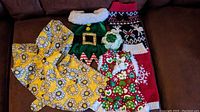 Assortment of five small dog clothing items including raincoat, elf costume, holiday sweater, festive dress, and fluffy collar.