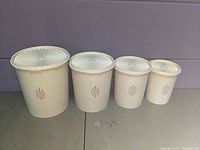 Four white vintage Tupperware canisters in graduated sizes lined up side by side showing the pink floral design and starburst lids.