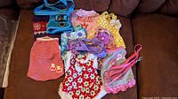 Photo showing assorted XXS dog sweaters, floral and Christmas dresses, and various harnesses and leashes arranged on a brown couch.