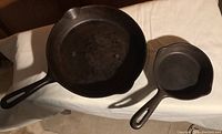Photo showing both cast iron fry pans side by side on a surface, one larger and one smaller, black finish, both with a handle and pouring spouts.