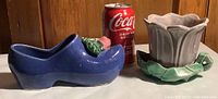 Blue shoe-shaped McCoy pottery planter with pink rose detail beside grey scalloped flowerpot with green leaf-shaped base, scale reference with Coca-Cola can.