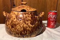 Front angle view of large bean pot next to a soda can for scale, showing mottled brown and tan glaze finish and knob handle on lid.
