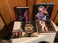 Photo of six music books including Jimi Hendrix, Bruce Springsteen, Pink Floyd, Bob Dylan, and Janis Joplin arranged on a white cloth against a wooden wall.