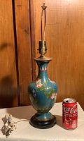 Front view of cloisonné lamp base next to a Coca-Cola can for scale, showing turquoise blue enamel with pink and yellow floral designs.