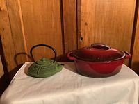 Photo of green dragonfly cast iron teapot and red Dutch oven on a white cloth-covered table with wood panel background.