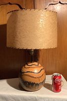 Full view of lamp with lava glaze pottery base and beige textured lampshade beside a can for size reference.