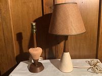 Two mid century table lamps on a table against wood paneling. One lamp has a ceramic pottery base with tapered shape and a tan fabric conical shade; the other has a rounded teak and wood base and no shade.