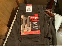 Two pairs of Wrangler Riggs Workwear pants with tags displaying size 32x34 and features like Room2Move Comfort, tough durable construction, patented tape measure reinforcement, side cargo pockets, and lifetime warranty.