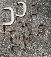 Photo of 7 assorted C-clamps on asphalt surface showing variety of sizes and brands including Williams, Armstrong, and malleable clamps.