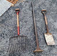 Photo of a hay and potato pitch fork with wooden handle and red metal head, a metal hand tamper with long shaft, and a shovel with wooden handle and metal blade, all placed on blacktop surface.