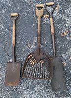 Three vintage garden tools laid flat on pavement: a rectangular shovel, a wide rake-like shovel tool, and a pointed spade shovel, all with wooden handles and metal working ends showing age and wear.