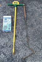 Overall image of three garden tools on pavement: yellow Dig It weed remover, boxed folding shovel, metal weedeater with wooden handle