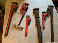 Full view of six assorted pipe wrenches and one pipe cutter laid on a light-colored surface, showing overall size and condition.
