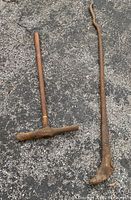 Two vintage railroad tools lying on asphalt: one is a spike driver with a wooden T-handle and long shaft, the other is a large rusted pry bar with a hooked end.