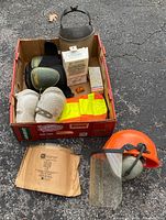 Photo showing box containing two sets of knee pads (black and white), bright yellow fabric pieces, and an orange hard hat with clear face shield and earmuffs with a partially broken strap.