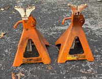 Two orange 2-6 ton automotive jack stands positioned on pavement, showing the bases, locking mechanisms, and saddles with rust and paint wear.
