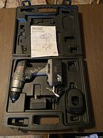 Open black plastic carrying case showing cordless drill with battery attached, charger, and instruction manual inside case molded compartments.