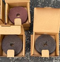 Photo of three opened boxes showing sanding discs inside. Discs are dark colored, round with central hole for sanding tool attachment.