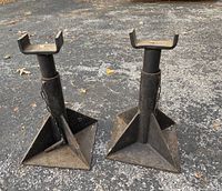 Photo showing two vintage metal jack stands with wide base and U-shaped saddle tops placed on asphalt surface
