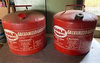 Photo showing two red vintage galvanized steel gas cans with large white text and eagle logo, metal handles on top, one with red cap, one with black cap, placed side by side on a wooden surface.