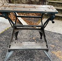 Side-front angled view showing overall structure, wooden top platform with holes, clamping handle on right, pedal/platform at bottom, steel frame and legs with wear and some rust.