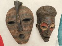 Side by side view of two African wooden masks, large mask with beaded mouth on left and smaller mask with painted eyes on right.