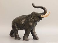 Front left angle view of bronzed elephant sculpture with raised trunk and two light-colored tusks, highlighting detailed skin folds and ears.