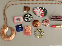 Photo showing all jewelry items laid out: a large textured copper crescent necklace, several colorful enamel brooches/pins, and a lipstick-shaped brooch.